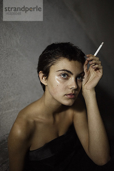 Schöne junge Frau schaut weg  während sie eine Zigarette gegen eine graue Wand hält