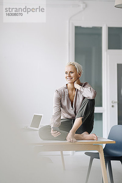 Nachdenkliche Unternehmerin mit Smartphone auf dem Schreibtisch vor der Wand im Büro