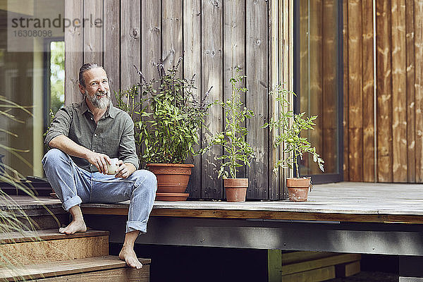 Lächelnder bärtiger Mann hält Kaffeetasse  während er vor einem kleinen Haus sitzt