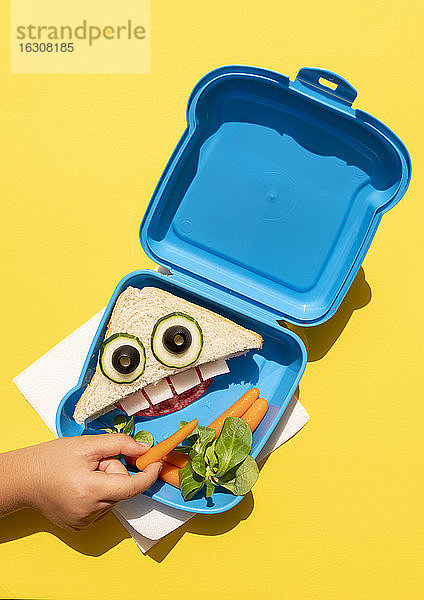 Hand von Baby-Mädchen abholen Baby Karotte aus Lunch-Box mit lustig aussehenden Sandwich mit anthropomorphen Gesicht