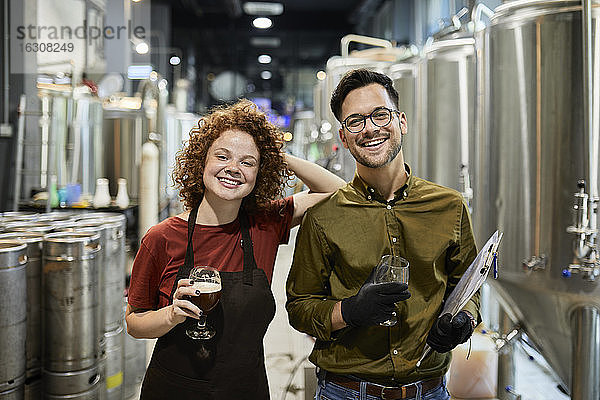 Porträt eines glücklichen Mannes und einer Frau  die ein Klemmbrett und ein Bierglas in einer Handwerksbrauerei halten