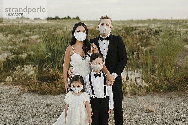 Eltern und Kinder in Hochzeitskleidern tragen einen Mundschutz  während sie während COVID-19 im Feld stehen