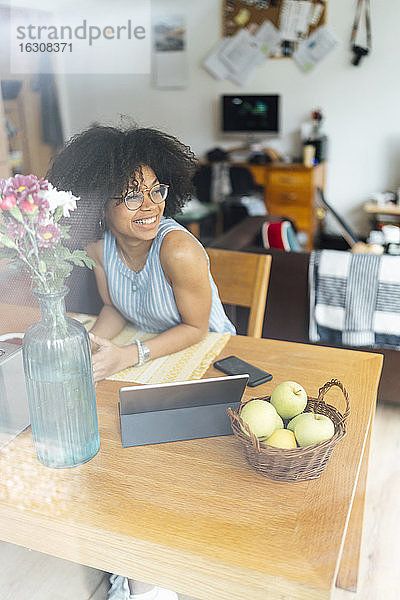 Lächelnde Geschäftsfrau mit digitalem Tablet auf dem Schreibtisch  die wegschaut  während sie zu Hause sitzt