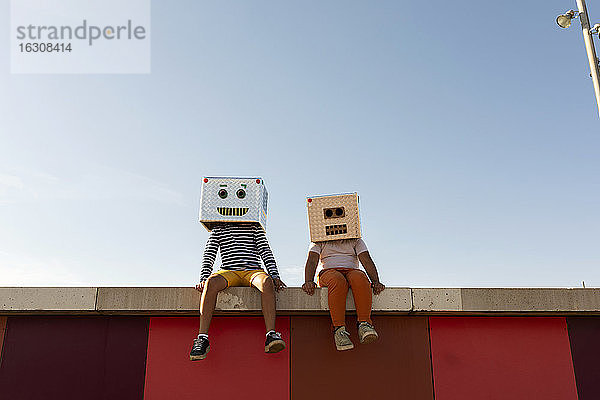 Freunde mit Robotermasken sitzen auf einer Stützmauer vor einem strahlend blauen Himmel