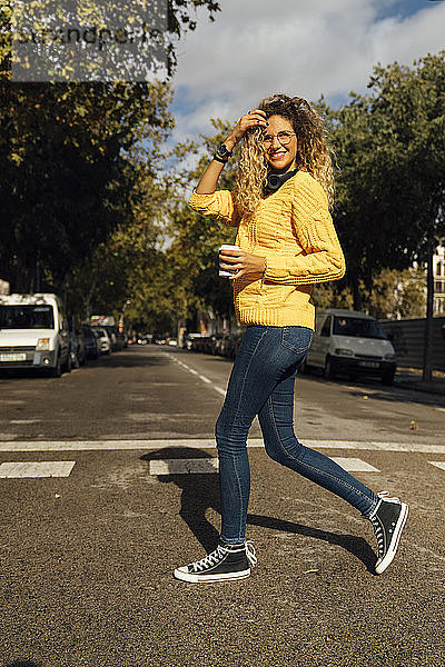 Lächelnde junge Frau beim Überqueren der Straße mit einem Einwegbecher in der Stadt an einem sonnigen Tag