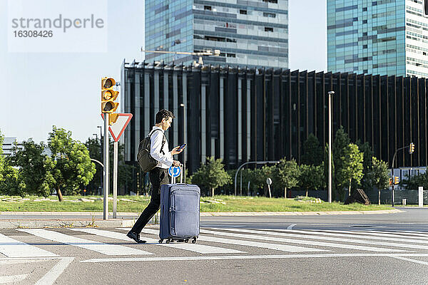 Geschäftsmann mit Koffer benutzt Smartphone beim Überqueren der Straße in der Stadt