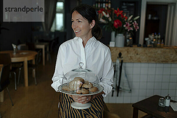 Lächelnde Besitzerin hält Kekse in einem Teller mit Cloche in einem Cafe