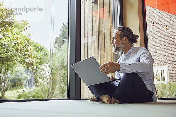 Bärtiger reifer Mann mit Laptop schaut durch das Fenster  während er in einem kleinen Haus sitzt