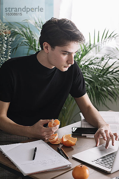 Junger Mann  der frische Orangenfrüchte isst  während er einen Laptop benutzt und Hausaufgaben macht