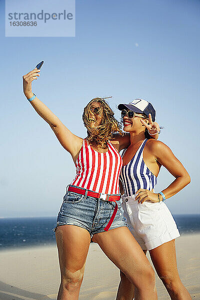 Fröhliche Freundinnen  die ein Selfie machen  während sie am Strand gegen den klaren Himmel stehen