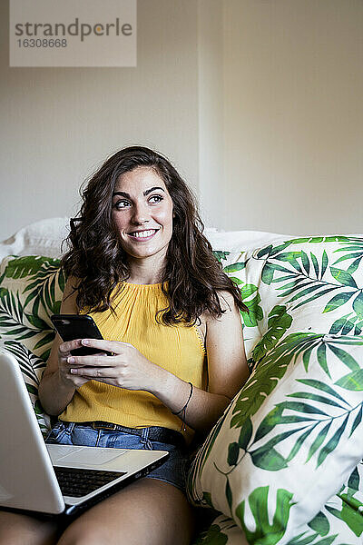 Nachdenklich lächelnde Frau  die ihr Handy in der Hand hält  während sie mit ihrem Laptop auf einem Sessel zu Hause sitzt