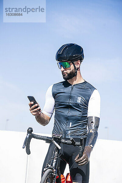 Männlicher amputierter Radfahrer  der sein Smartphone benutzt  während er mit dem Fahrrad vor einem klaren Himmel steht