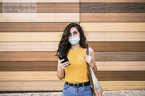 Junge Frau hält Smartphone und Netztasche  während sie an einer Holzwand steht