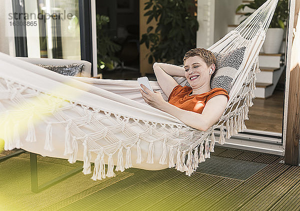 Lächelnde Frau mit drahtlosen Kopfhörern  die ihr Smartphone benutzt  während sie auf einer Hängematte auf der Veranda liegt