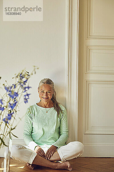 Lächelnde Frau mit gekreuzten Beinen auf dem Boden sitzend gegen die Wand zu Hause