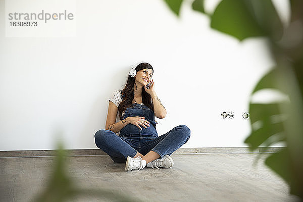 Lächelnde schwangere Frau hört Musik über Kopfhörer  während sie in ihrem neuen Haus an der Wand sitzt