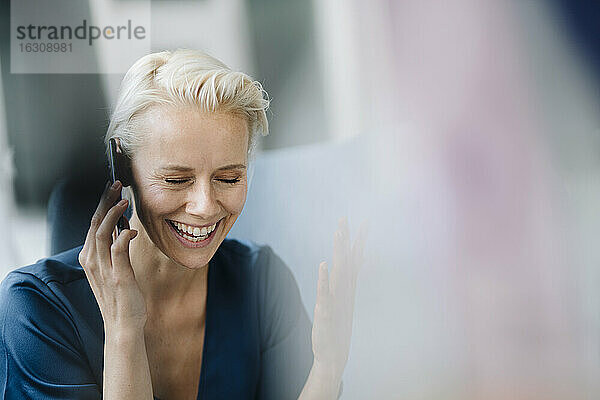 Nahaufnahme einer fröhlichen Geschäftsfrau mit geschlossenen Augen  die über ein Smartphone im Büro spricht