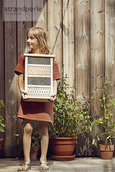 Mädchen  das ein Insektenhotel trägt  während es im Hof an der Wand steht