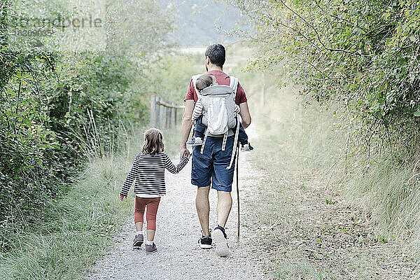 Vater wandert mit seinen Kindern  trägt seinen Sohn auf dem Rücken und hält die Hand seiner Tochter