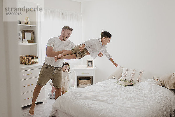 Verspielter Vater  der seinen Sohn über das Bett wirft  während die Tochter im Schlafzimmer steht