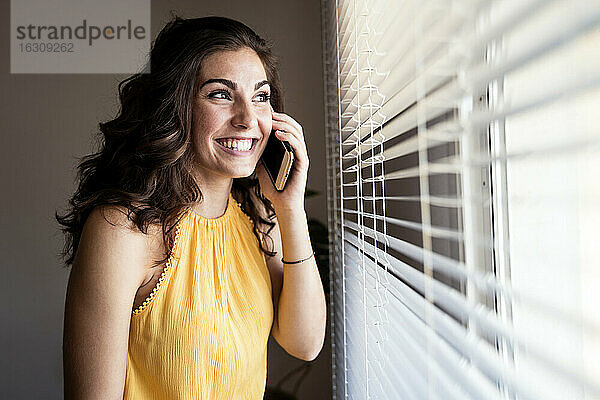 Fröhliche junge Frau  die mit ihrem Smartphone spricht  während sie durch die Jalousien zu Hause wegschaut