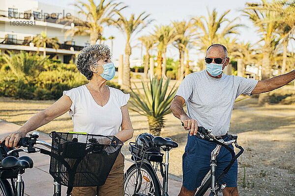 Älteres Paar mit Gesichtsmaske  das mit dem Fahrrad im Park spazieren geht