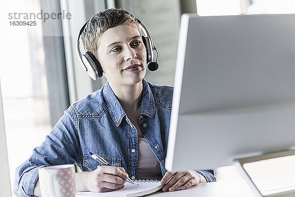 Geschäftsfrau mit Headset am Schreibtisch im Büro macht sich Notizen