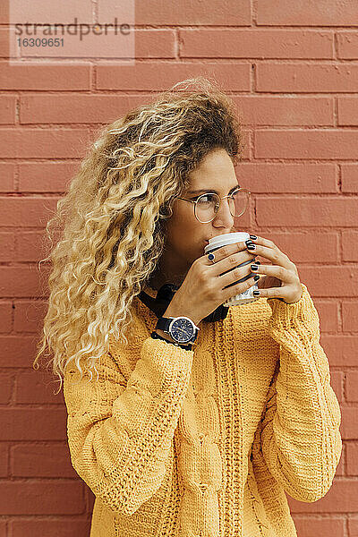 Junge Frau trinkt Kaffee aus einem Einwegbecher  während sie gegen eine rote Backsteinmauer schaut