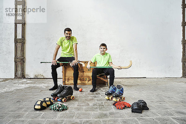 Vater und Sohn sitzen mit Hockeyschlägern auf einer Holzkiste bei den Sportgeräten an der Wand im Gericht