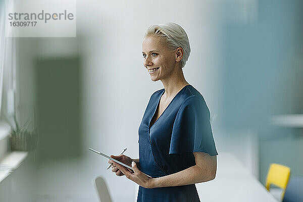 Lächelnde Geschäftsfrau mit digitalem Tablet  die wegschaut  während sie im Büro steht