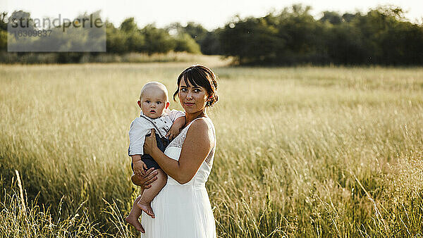 Die Braut trägt einen kleinen Jungen  während sie bei Sonnenuntergang auf einem Feld steht