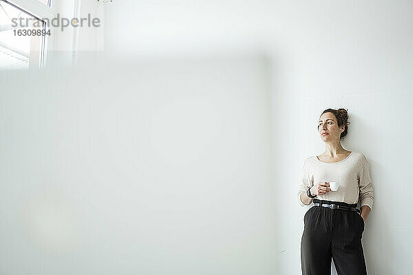 Geschäftsfrau  die eine Kaffeetasse hält und nachdenklich vor einer weißen Wand steht
