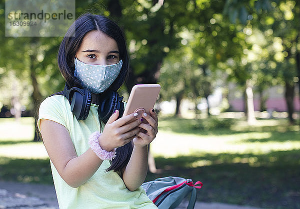 Mädchen mit Gesichtsmaske benutzt Smartphone in öffentlichem Park