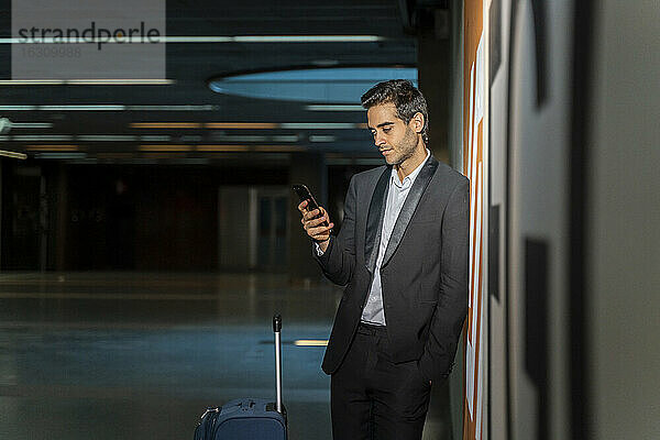 Männlicher Berufstätiger  der ein Mobiltelefon benutzt  während er mit einem Koffer an der Wand eines Bahnhofs steht