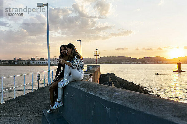 Freunde telefonieren  während sie bei Sonnenuntergang auf einer Stützmauer an der Promenade sitzen