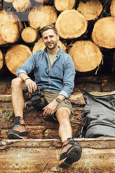 Lächelnder Mann mit Fernglas und Rucksack sitzt auf einem Baumstamm vor einem Holzstapel
