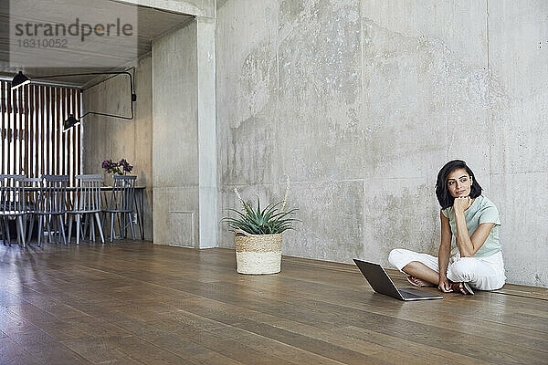 Nachdenkliche Geschäftsfrau sitzt mit Laptop auf Hartholzboden vor der Wand im Büro