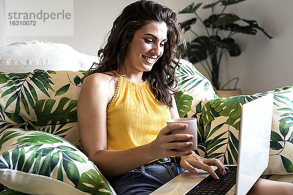 Lächelnde schöne Frau  die eine Kaffeetasse hält und einen Laptop benutzt  während sie zu Hause auf einem Sessel sitzt