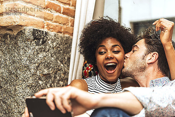 Nahaufnahme eines Mannes  der seine fröhliche Freundin küsst  während er ein Selfie mit ihr in einem Café macht