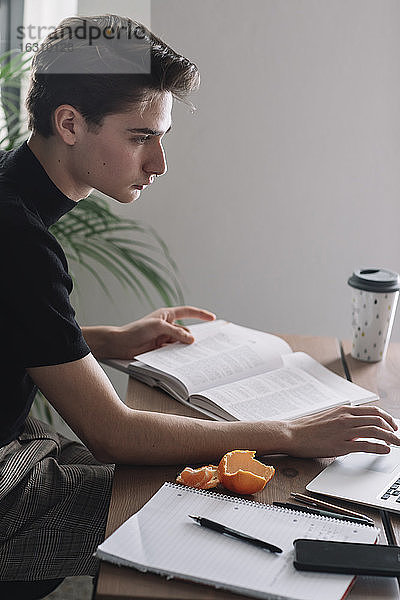 Gutaussehender männlicher Student  der einen Laptop benutzt  um am Tisch Hausaufgaben zu machen