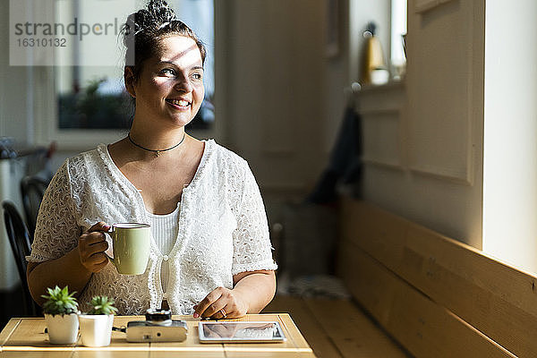 Junge Frau hält Kaffeebecher nachdenklich  während sie am Tisch in einem Café sitzt