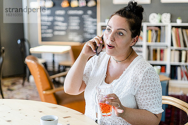 Üppige Frau  die ein Getränk hält und über ein Mobiltelefon spricht  während sie im Restaurant sitzt