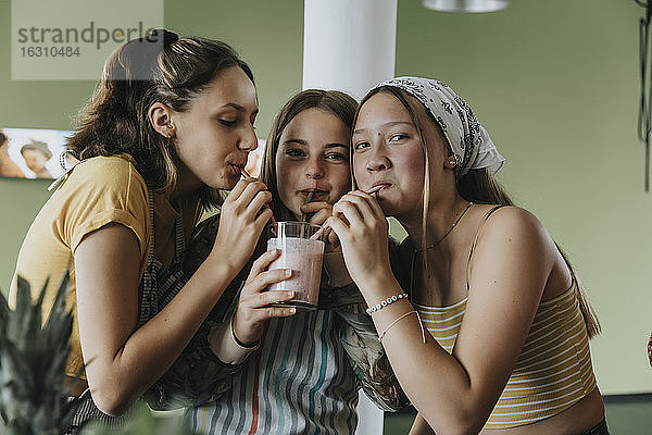Teenager-Mädchen stehen in der Küche und teilen sich einen frischen Frucht-Smoothie mit Trinkhalmen