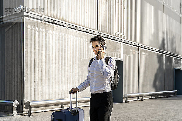 Geschäftsmann mit Koffer  der über sein Smartphone spricht  während er auf dem Fußweg vor einem Gebäude steht