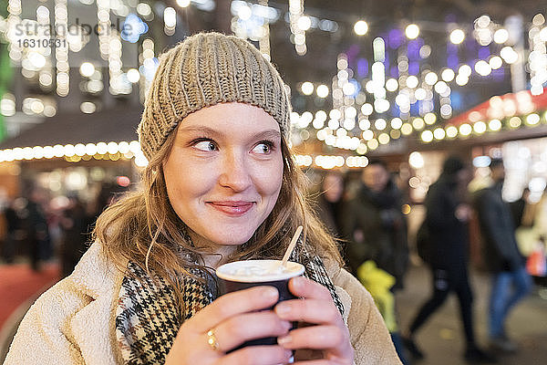 Nahaufnahme einer schönen Frau mit heißer Schokolade auf dem Weihnachtsmarkt bei Nacht