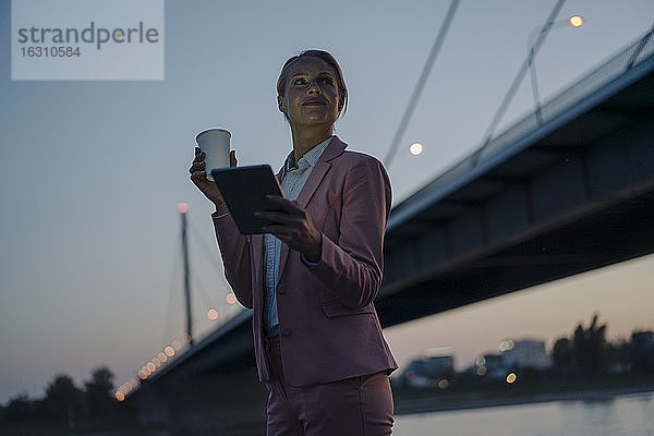Geschäftsfrau schaut weg  während sie ein digitales Tablet und einen Einwegbecher gegen eine Brücke in der Abenddämmerung hält