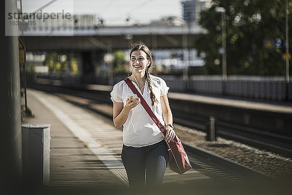 Lächelnde Frau hört Musik über ihr Mobiltelefon  während sie auf dem Bahnhof spazieren geht