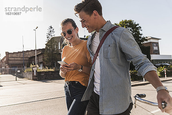 Lächelndes junges Paar  das auf sein Handy schaut  während es auf der Straße in der Stadt spazieren geht