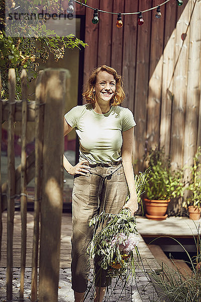 Lächelnde erwachsene Frau  die Blumen hält  während sie vor einem kleinen Haus steht