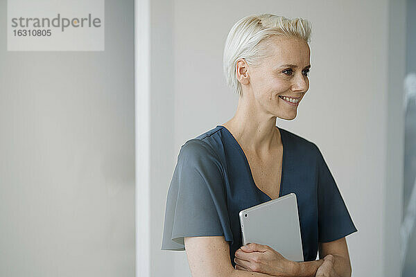 Lächelnde Geschäftsfrau  die ein digitales Tablet hält und wegschaut  während sie im Büro an der Wand steht